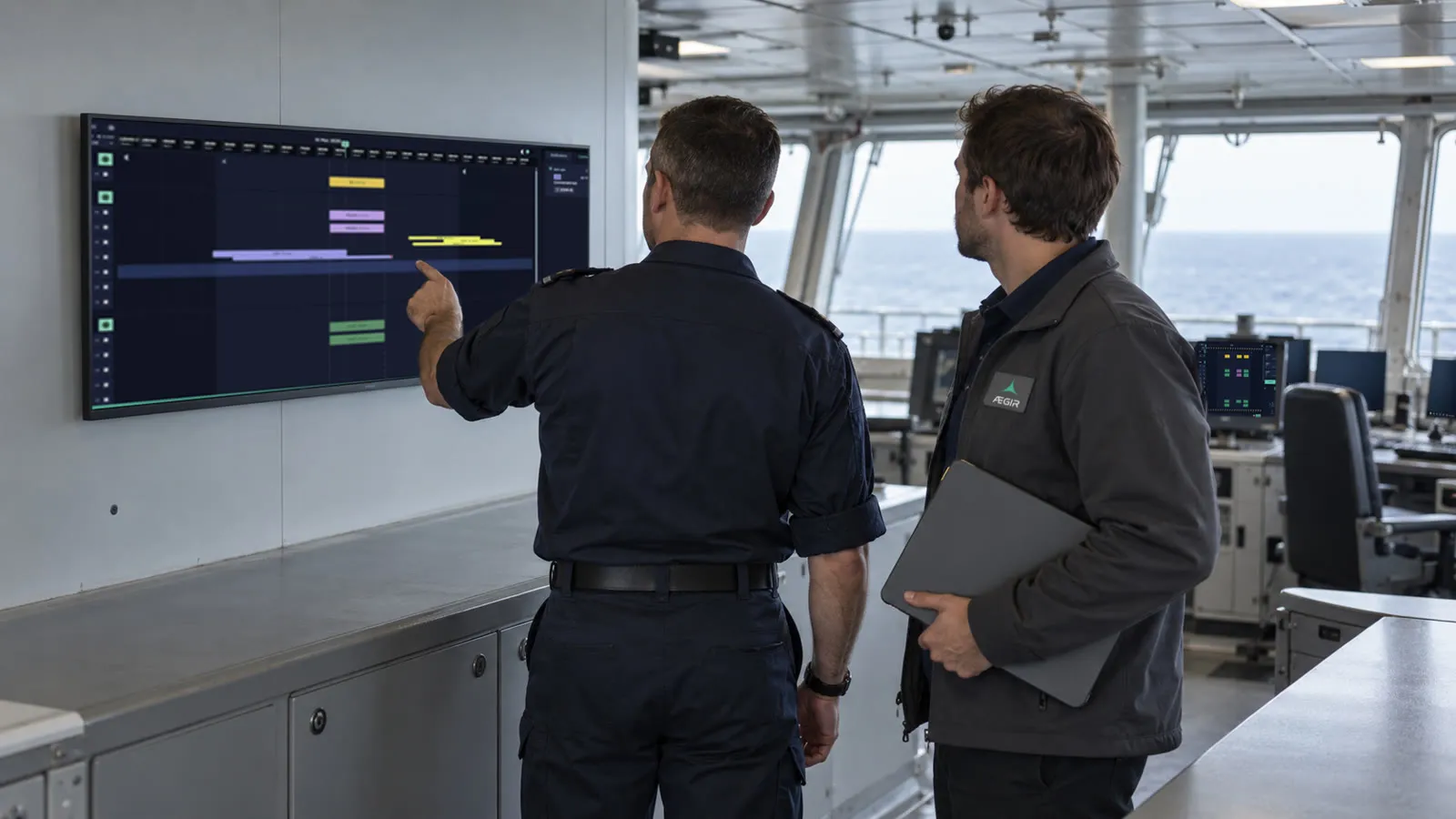 On a ship's bridge by day, a naval officer in a dark uniform, seen from behind, points at a large wall-mounted display showing a planning timeline, while an AEGIR engineer holding a laptop looks on; workstations and the open sea are visible behind them.