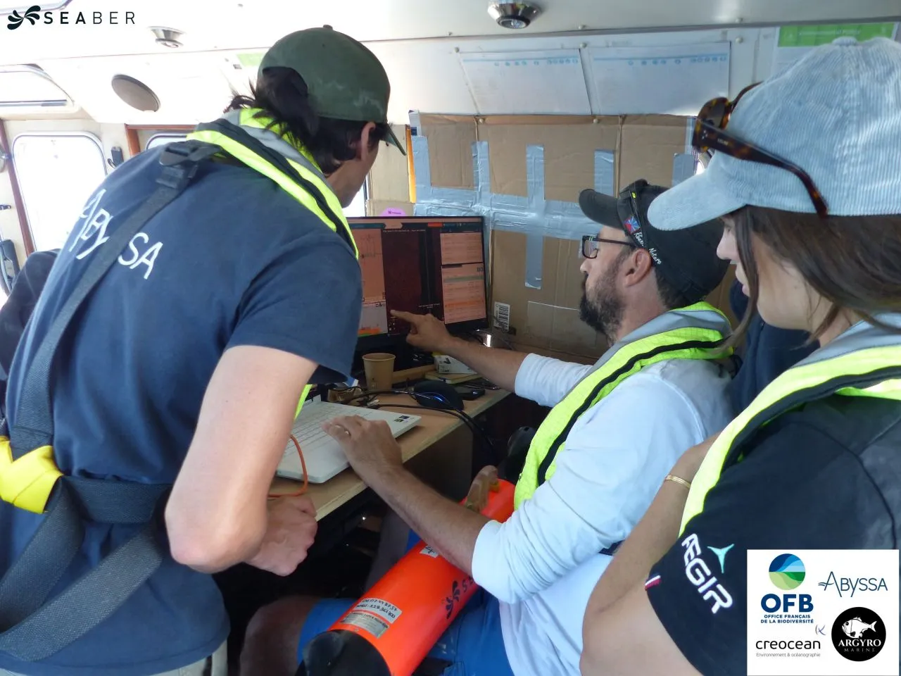Members of the ABYSSA and AEGIR teams in high-visibility vests gathered around a screen during the underwater exploration campaign.